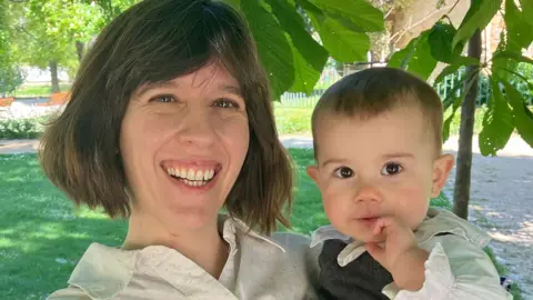 BBC Tania has brown hair and is holding her baby in a sunny park.