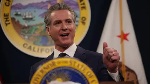 San Francisco Chronicle via Getty Images California Governor Gavin Newsom speaks at a podium bearing the California state seal. The California flag, which features a grizzly bear and a red star, is visible in the background.