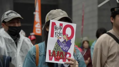 BBC News: Jiro Akiba A person holds a sign over their face with the words 'NO WAR'
