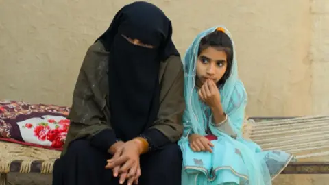 BBC A woman and a girl sit on a bench made of metal and woven fabric. The woman wears a dark veil that covers her face and head, with her eyes visible, while the girl is wearing light blue clothes and a scarf over the top of her head