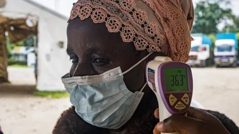 AFP via Getty Images A person wearing a mask and peach headwrap is having their temperature checked with a contactless thermometer near their ear. it reads 36.3 degrees.