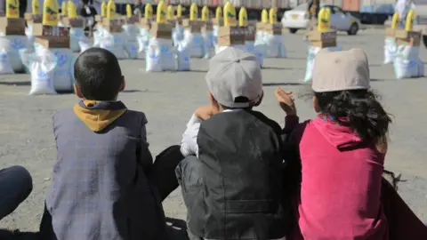 BBC Three children sitting, with their backs to the camera, facing bags of aid