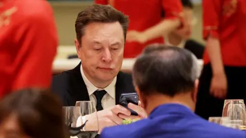 Alex Wong/Getty Images A man in a suit and tie stares at his phone while sitting down opposite a man in a blue jacket in Beijing