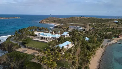 Getty Images Aerial view of Little St James island there is a large estate with multiple buildings, palm trees, and blue-roofed villas surrounded by the sea.