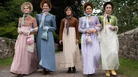 BBC/Bad Wolf/James Pardon The two bennet sisters on the left and right wear colourful period costumes of dresses and cropped cardigan ( L-R- pink, blue, purple, green). Mary, in the middle, wears a more basic white dress, a brown cropped cardigan and a beige head scarf.