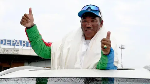 AFP via Getty Images Kami Rita Sherpa in a green winter jacket and a cap, gives the thumbs-up sign while standing in a car.