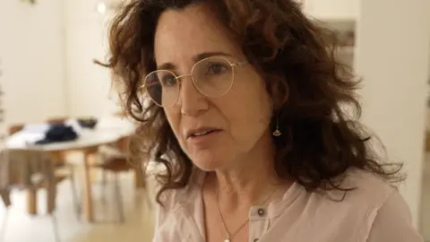 BBC Liat Zvi is pictured from the shoulders up, in a domestic setting with a table in the background. She is a dark-haired woman with gold-rimmed glasses.