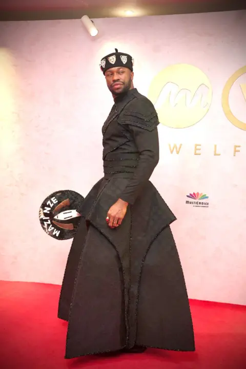 BBC/GIFT UFUOMA Nigerian actor Stan Nze is styled in a black apparel and carried his signature tradtional engraved hand fan made with horse skin.