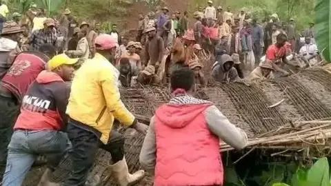 South Ethiopia Regional State Office Of The President A large group of people work together to clear or lift sections of a collapsed structure made from wooden poles and thatch. Many individuals stand on or around the debris, while others gather on a slope in the background. The scene is set in a green area with tall trees, dense vegetation and plants surrounding the site.