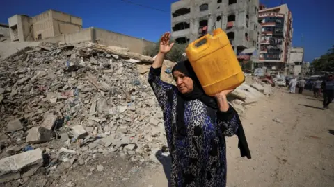 Reuters Palestinian woman carries a container of water as she passes by rubble in Gaza City (03/07/25)