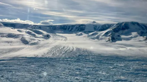 Naomi Ochwat Hektoria Glacier flows into the partly frozen ocean. The glacier surface is white with some cracks. The partly frozen ocean is light blue. In the background is a mountain range, sweeping round to the left as we look at it, covered in snow. The sky is lightly clouded with some Sun rays visible. 