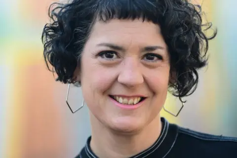 Laura Murphy Laura Murphy, a young woman with black curly hair and octagonal silver earrings, smiles at the camera against a neutral, light blue background