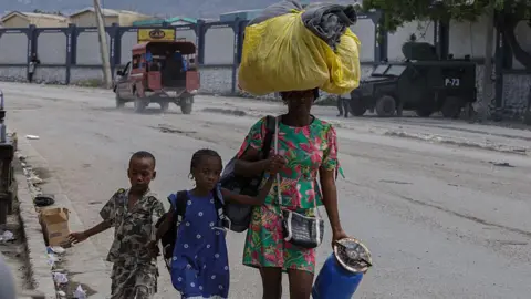 Getty Images A woman holds a canister and has a bag on her head. She wears a green dress. In her other hand she holds the hand of a girl. The girl holds the hand of a boy. 