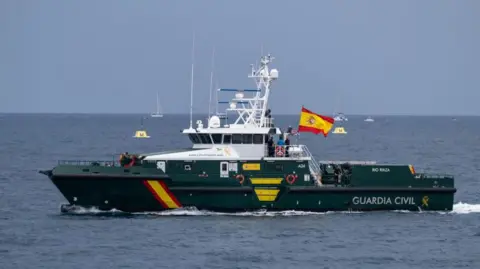 Getty Images A medium-sized green boat with the works 'Guardia Civil' on the side sails in the ocean. There are a few silhouettes of smaller boats in the far distance.
