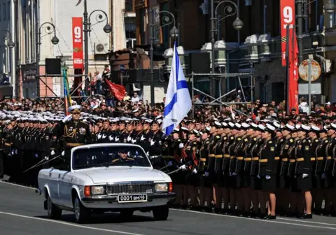 Reuters A Russian military commander salutes while standing in an open-top, white car being driven by another soldier on a road flanked by rows of military personnel. 