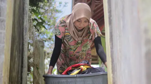 Getty Images Young Asian Muslim woman wearing a hijab and casual daily home outfit holds a plastic laundry basket filled with clothes in the morning