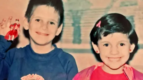 Alex Castilleja A boy in a blue top and a girl in a pink top, both with dark hair, smile at the camera - the boy is holding up a toy figurine 