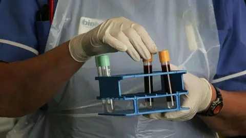 Getty Images A Biobank employee wearing a blue nurse's uniform, white apron and white disposable gloves, handles vials of blood in a lab.