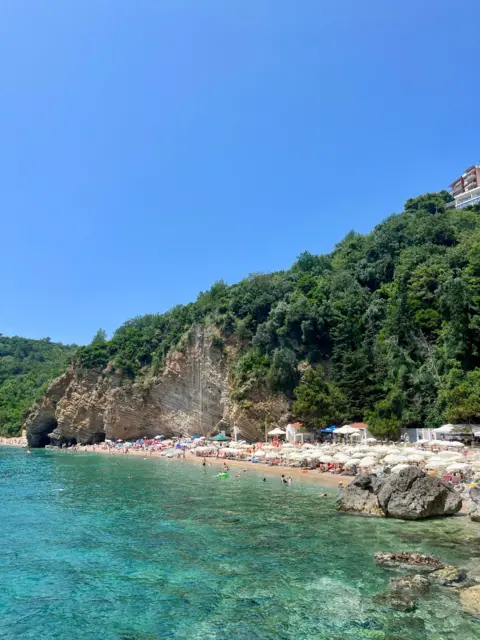 Em's Budget Travel Landscape image of a beach in Montenegro. There is a large, high landmass jutting out into the sea. The water is clear blue and the sky is clear and sunny.