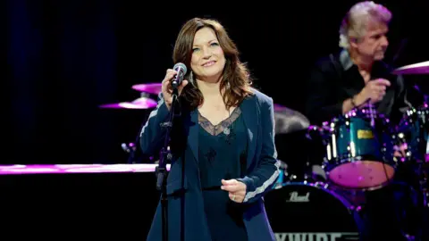 Getty Images A woman with wavy brown hair in a black pant suit smiles in front of a microphone 