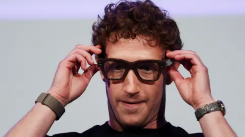 Reuters Meta CEO Mark Zuckerberg on a stage motioning toward the thick framed eye glasses on his face which are that are a prototype of the company's unreleased AI-powered smart glasses. He is wearing a black t-shirt and a wrist watch.