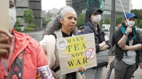 BBC News: Jiro Akiba A woman with grey hair holds a sign that says 'Make tea not war'