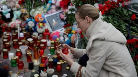 Reuters A woman with blonde hair tied back, wearing a cream coat and gold-rimmed glasses lights a candle at a makeshift memorial in Krasnogorsk, outside Moscow.