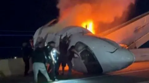 ZAYRA GARZA Wreckage of jet and multiple bystanders are in the image, helping pull people from the plane. 
