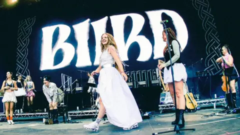 Kate Lawlor Members of BIIRD perform on an outdoor stage in front of a large screen displaying the band’s name, with musicians in varied outfits standing and moving across the stage alongside instruments and microphones.
