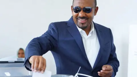Reuters Abiy Ahmed in a navy suit, white shirt and aviator glasses smiles as he places a ballot in a ballot box. Behind him a female electoral official can be seen.