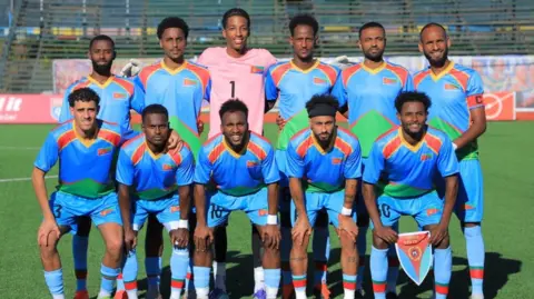 ENFF A team photo posed before a match by the Eritrean men's football side. The outfield players are wearing all-blue kit with red, green and gold trim. Five players in the front row bend forward with their hands placed on their knees, with the player on the far right of the front row holding a team pennant. Six players stand in the back row with their arms wrapped around the team-mate to either side. The goalkeeper is in a pink jersey with the number one on its chest. The players are on a green pitch with an empty wooden stand in the background