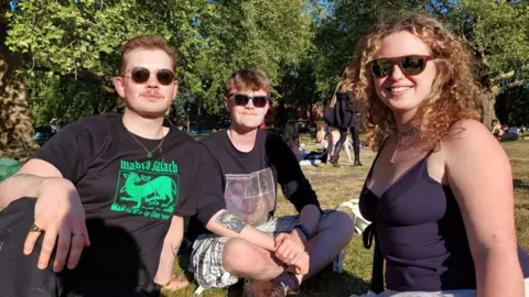 Three friends - two men and a woman - sit on the grass in a park in sunglasses, smiling