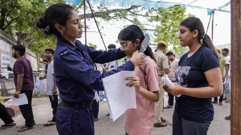 Himanshu SHARMA / AFP via Getty Images A female security woman is running a hand-held metal detector over a young woman's hair, as others wait in line.