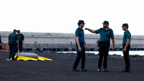 Reuters Three men in police uniform stand at the right of the picture, while two others stand further back on the left over a grey and yellow tarpaulin or canvas, as they prepare the port of Grenadilla for the arrival of the MV Hondius