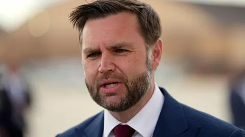 Getty Images A close-up image of JD Vance talking to reporters. He wears a dark blue jacket, white shirt and dark red tie. He has short brown hair.