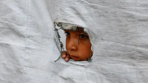 Reuters A displaced Palestinian boy peers through a tear in a tent, in Gaza City, northern Gaza (6 May 2026)