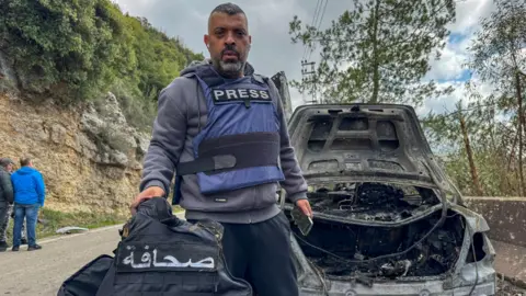 Reuters A man in a press vest stands beside a blown-out car on a hillside, holding a press vest towards the camera