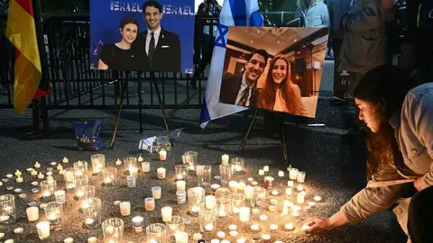 AFP via Getty Images dozens of lit candles on the sidewalk in front of two posterboards displaying photos of both the victims