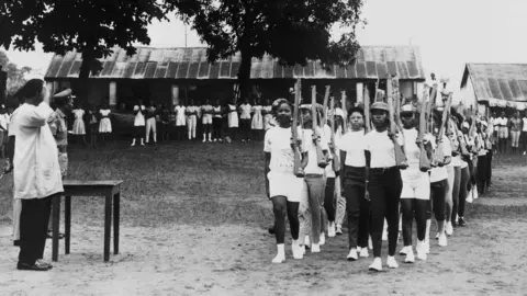 AFP via Getty Images Young female militia of the civil defence - most dressed in white and with white socks and trainers - hold what look like rifles against their shoulders as they parade during military training in Enugu in August 1967. To the side a man in a white coat and dark trousers salutes as two other soldiers look on. It is a black and white archive photo.