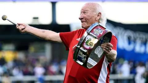 Getty Images An elderly man with long white hair wears a red-and-white sports shirt and bashes a drum he is holding in his left hand, whilst smiling and looking up at a crowd in a sports stadium.