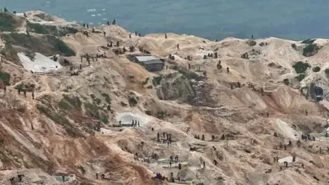 Hassan Lali / BBC Overhead shot of mining activities at Rubaya mine where the terrain is hilly - slightly terraced. The bare earth is a light brown, some of it has a darker more orange shade. Many groups of miners can be seen from a distance cleaning ore. One corrugated metal shack can be seen. In the distance are green fields.