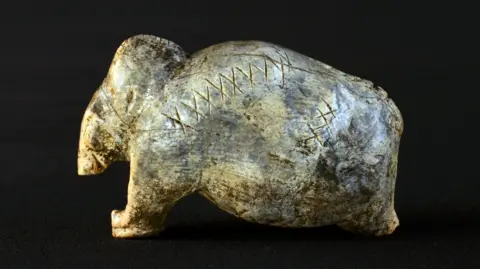 University of Tübingen / Hildegard Jensen A figurine shaped like a mammoth. It is ivory colour with dark grey patches, and has deeply-engraved lines of crosses and dots