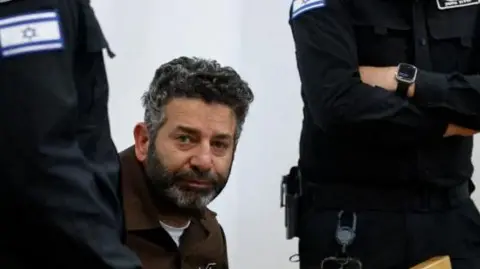 Reuters Saif Abu Keshek sits at a desk accompanied by two standing Israeli police officers at a court in Israel. He has short, greying hair and a beard.