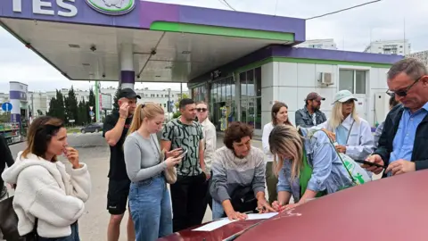 Reuters People dressed in casual clothing at a petrol station in Sevastopol, Crimea leaning on a car bonnet putting their names on a list to join the queue for fuel after the authorities restricted fuel sales amid a supply shortage following Ukrainian attacks on logistics routes in the course of Russia-Ukraine conflict, June 1, 2026.