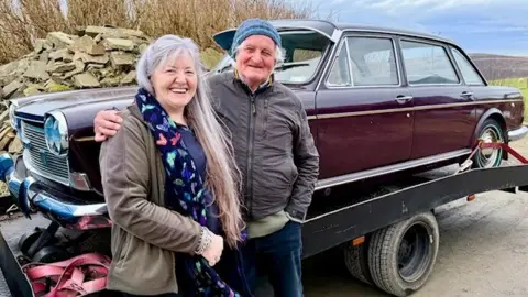 BBC A woman with long grey hair stands next to a man with a blue woollen hat. They are standing in front of an old brown car, which is on the back of a lorry. 