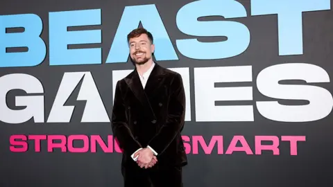 Getty Images MrBeast at the premiere of Beast Games, which is written in white against a black background behind him. He is wearing a black blazer and trouser and white shirt.