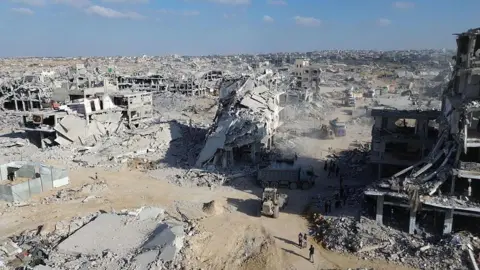 Anadolu via Getty Images A picture from 28 October, 2025 shows a truck and four other vehicles surrounded by rubble in Khan Younis. All the buildings in the foreground and middle-distance have been totally collapsed into dust and debris. 