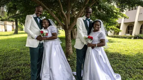 AFP via Getty Images The couples pose for a photo. The brides wear identical white dresses while the grooms wear matching suits and bow ties. 