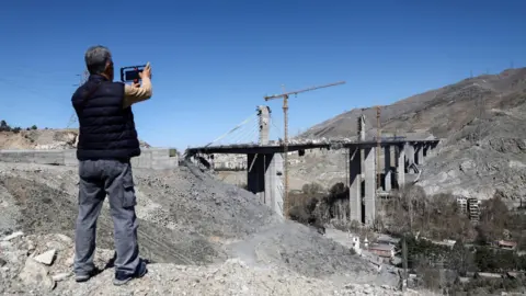WANA via REUTERS A man uses his phone to photograph the remains of the B1 bridge following a US air strike, in Karaj, northern Iran (3 April 2026)