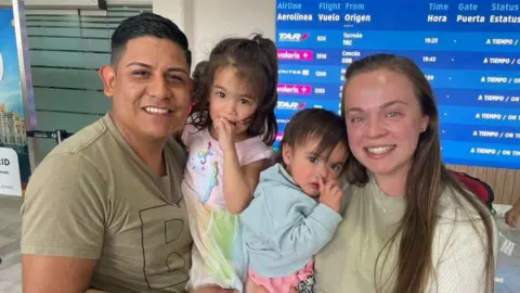 Submitted by Janie Pérez Janie and Alejandro smile, hold their two daughters in front of a boarding flights sign